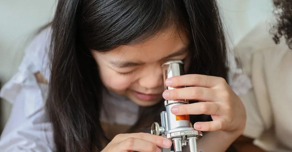 Cute little Asian girl with long dark hair making experiment with microscope while sitting at wooden table in classroom