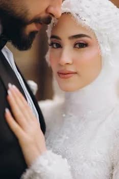 Close-up portrait of a wedding couple with bride in a white hijab embracing groom in a tuxedo.