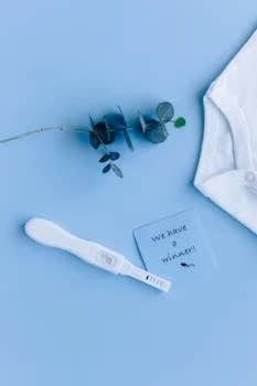 Flat lay of positive pregnancy test with baby onesie and note on blue background.