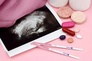 A closeup of pregnancy tests and an ultrasound with supplements on a pink background.