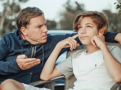 Father expressing frustration while teenage son shows defiance, seated outdoors.