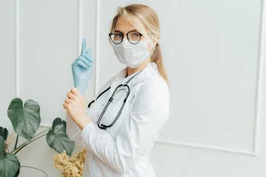 Healthcare professional in a white coat wearing a face mask and gloves, holding a stethoscope.