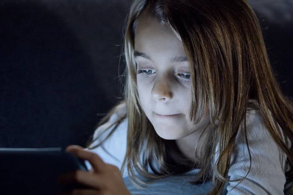 A young girl using a digital tablet in bed, bathed in soft light, focused on the screen.