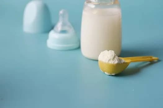 Close-up of baby milk formula powder in a yellow scoop with a bottle on a blue background.