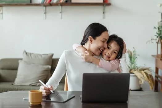 Happy young Asian woman working remotely from home with laptop and tablet while adorable little daughter hugging from behind