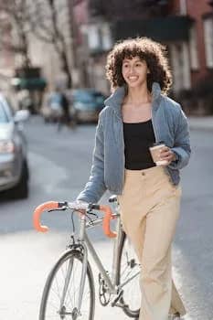 A cheerful young woman walks her bicycle while enjoying a coffee cup in an urban setting, embracing a lively lifestyle.