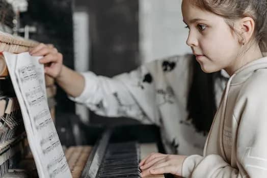 Girl in hoodie learning to play piano with sheet music during indoor lesson.